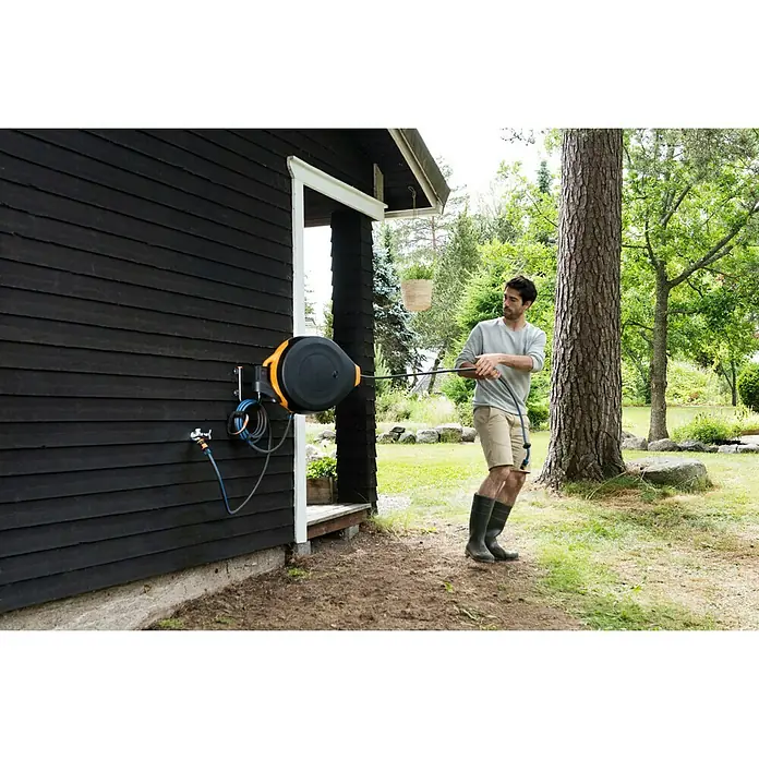 Fiskars Wand-Schlauchhalterung - Passend für: Fiskars Schlauchwagen Waterwheel XL - Ein Mann benutzt eine automatische Gartenschlauchbox an einer dunklen Holzwand, um seinen Garten zu bewässern.