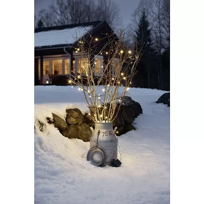 Konstsmide LED-Lichterkette - Außen, Lichtfarbe: Warmweiß, Batteriebetrieben, 0,5 m, Schwarz - Eine winterliche Außendekoration: Eine graue Metallmilchkanne mit kahlen Ästen, geschmückt mit warmweißen Lichterketten, solarbetrieben im Schnee.