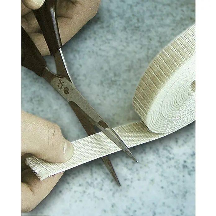 Schellenberg Cinta para persiana - Blanco, Largo: 6 m, Anchura de la correa: 23 mm - Un primer plano de una persona cortando una cinta de tela blanca con unas tijeras negras, con un rollo de cinta de tela al fondo.