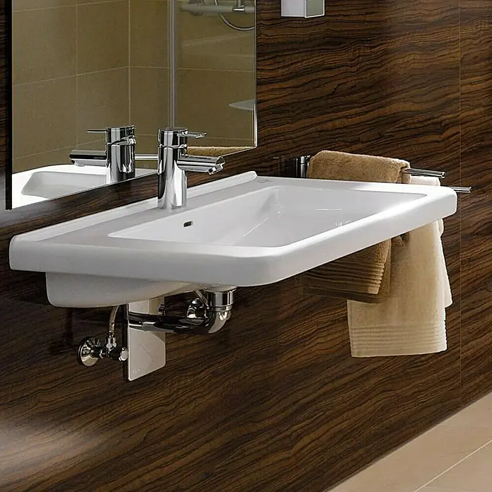 Geberit Renova Comfort Waschtisch - 55 x 52,5 cm, Mit schmutzabweisender Glasur, Hahnloch: Mitte, Mit Überlauf, Weiß - Das Bild zeigt ein modernes Waschbecken mit Armatur und Handtüchern vor einer Holzwand. Das Becken ist weiß und rechteckig.