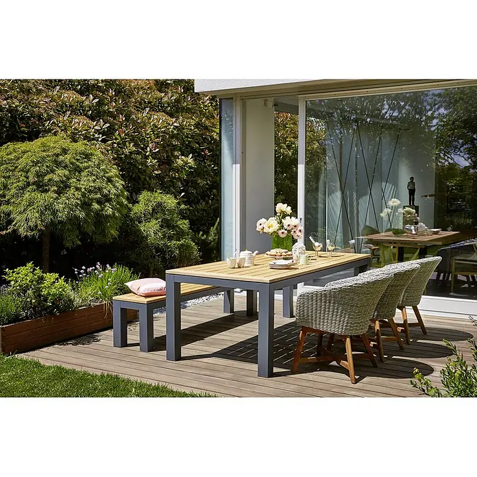 Sunfun Gartenbank Lynn - B x T x H: 195 x 40 x 45 cm, Naturbraun/Anthrazit - Ein elegantes Gartenset mit Tisch, Bank und Stühlen, perfekt für Mahlzeiten im Freien, präsentiert auf einer Holzterrasse mit üppigem Grün im Hintergrund.