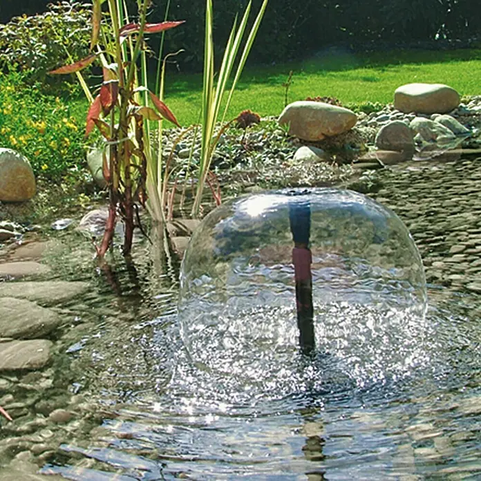 Neptun Premium Wasserspielpumpe NPTP-O 2500 - 40 W, Fördermenge pro Std.: 2.500 l - Eine Nahaufnahme eines sprudelnden Kugelbrunnenaufsatzes in einem Gartenteich mit Steinen, Pflanzen und Rasen im Hintergrund.