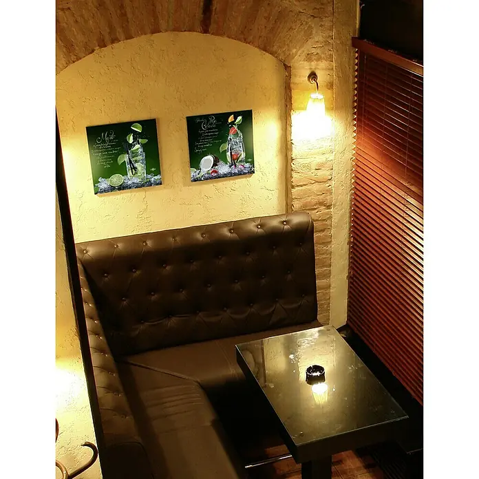 Glasbild - Cocktails II, B x H: 50 x 50 cm - Gemütliche Sitzecke mit brauner Ledercouch, einem Tisch mit Aschenbecher und Bildern von Cocktails an der Wand, beleuchtet durch eine Wandleuchte.