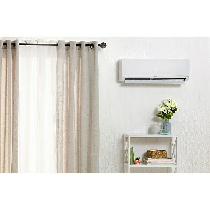 PR Klima Aire acondicionado Inverter PRO-18DH - 5.000 W, 5.300 W, 35 m², Blanco - Un aire acondicionado split blanco cuelga en una pared blanca sobre una estantería con decoraciones. Cortina beige frente al aparato.
