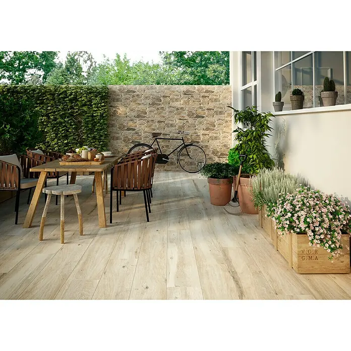 Halcón Pavimento porcelánico Atelier - 23,3 x 120 cm, Beige, Clase 2 - La imagen muestra un área al aire libre con baldosas de suelo con aspecto de madera, una mesa de comedor y una bicicleta contra una pared de piedra.