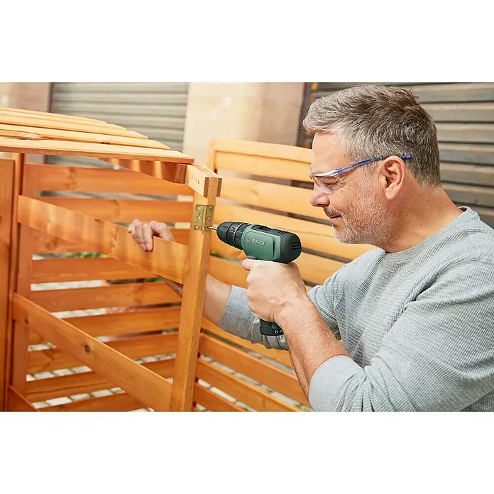Bosch Taladro atornillador percutor de batería EasyImpact 1200 - 12 V, 1 batería, 1,5 Ah, 30 Nm - Un hombre usa un taladro inalámbrico Bosch verde para conectar una bisagra a un cobertizo de madera, gafas de seguridad incluidas.