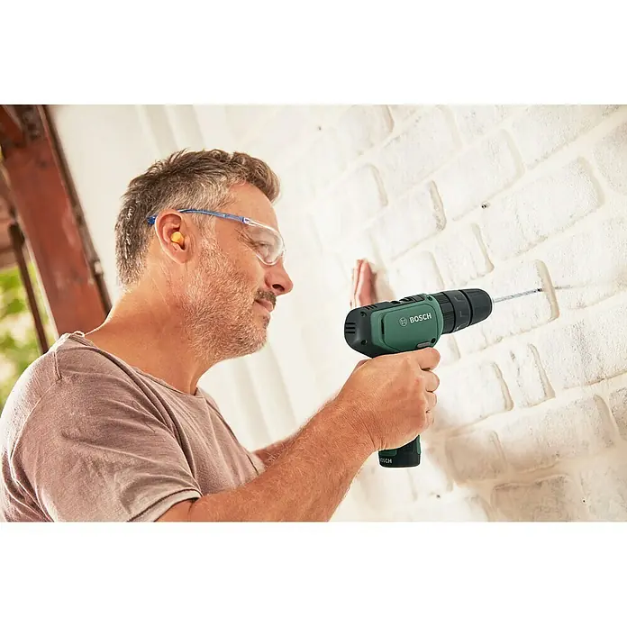 Bosch Taladro atornillador percutor de batería EasyImpact 1200 - 12 V, 1 batería, 1,5 Ah, 30 Nm - Un hombre taladrando una pared de ladrillo blanco con un taladro inalámbrico Bosch verde. Lleva gafas de seguridad y tapones para los oídos.