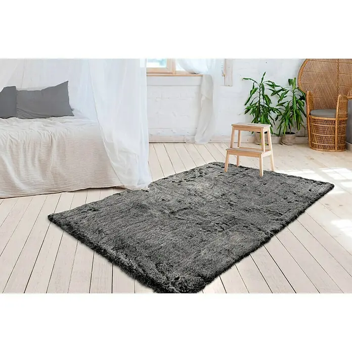 Kayoom Hochflorteppich Tender - Anthrazit, 170 x 120 cm - Ein grauer, flauschiger Teppich liegt auf einem Holzboden in einem hellen Raum mit einem Bett und Pflanzen.