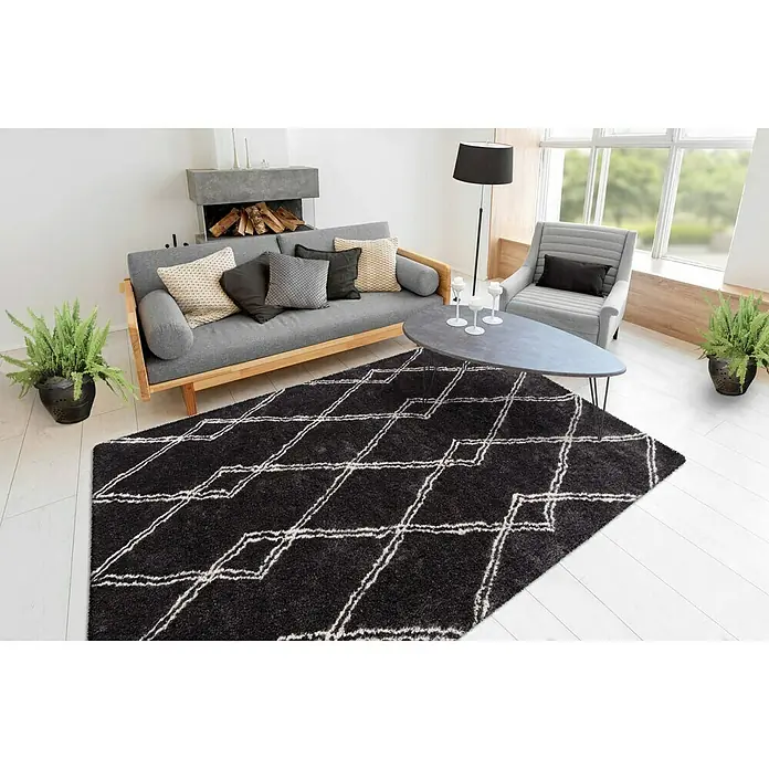 Kayoom Hochflorteppich Orlando 125 - Anthrazit, 230 x 160 cm - Ein stilvolles Wohnzimmer mit einem schwarzen Teppich mit geometrischem Muster als Blickfang. Die graue Couch und Sessel ergänzen das moderne Design.