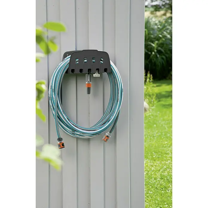 Gardena Soporte portamangueras de pared - Plástico, Apto para: Montaje en la pared - Una manguera de jardín Gardena en un soporte de manguera gris fijado a una pared, con un jardín verde al fondo.
