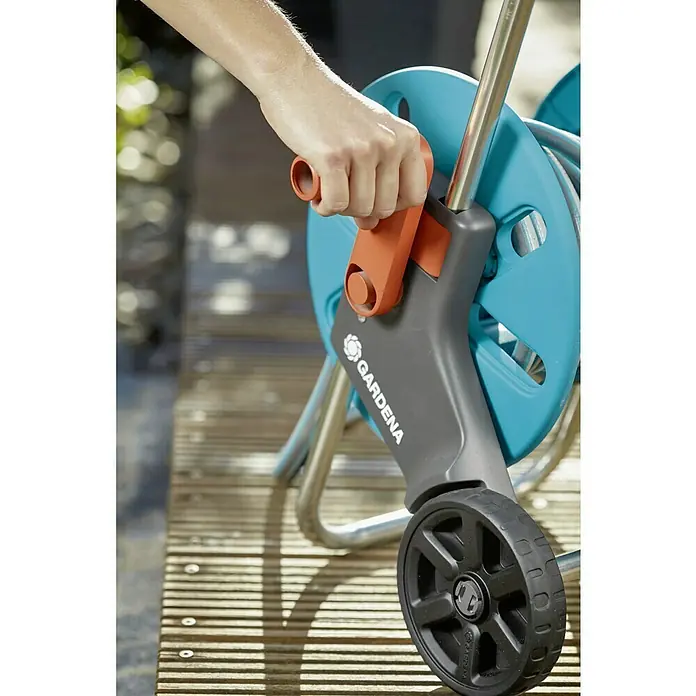 Gardena Kit carro portamangueras Aquaroll S - 20 m - Primer plano de una persona operando un carrete de manguera de jardín Gardena sobre una cubierta de madera, centrado en el asa y el logotipo de la marca.