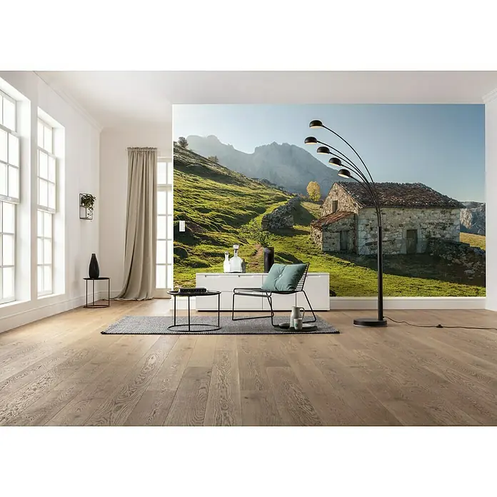 Komar Stefan Hefele Edition 2 Fototapete Picos De Europe Alm - 9 -tlg., B x H: 450 x 280 cm, Vlies - Ein elegantes Wohnzimmer mit Holzboden und einem Wandbild, das eine rustikale Berghütte zeigt. Die Lampe und der Sessel ergänzen das stilvolle Interieur.
