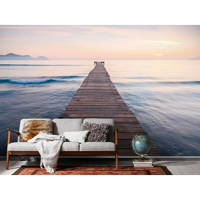 Komar Stefan Hefele Edition 1 Fototapete Peace - 4 -tlg., B x H: 400 x 250 cm, Vlies - Eine Couch mit Kissen und einer Decke vor einer Fototapete mit einem Steg ins Meer bei Sonnenuntergang, die eine entspannende Atmosphäre schafft.