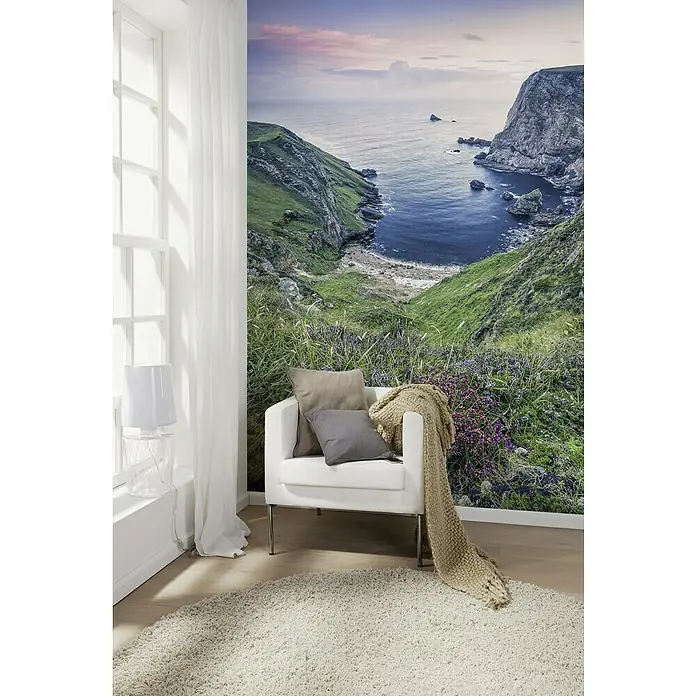 Komar Stefan Hefele Edition 1 Fototapete The Bluebay - 2 -tlg., B x H: 200 x 250 cm, Vlies - Die Nahaufnahme zeigt einen Sessel mit Kissen und Decke auf einem beigen Teppich vor einer Wandtapete mit Küstenmotiv.
