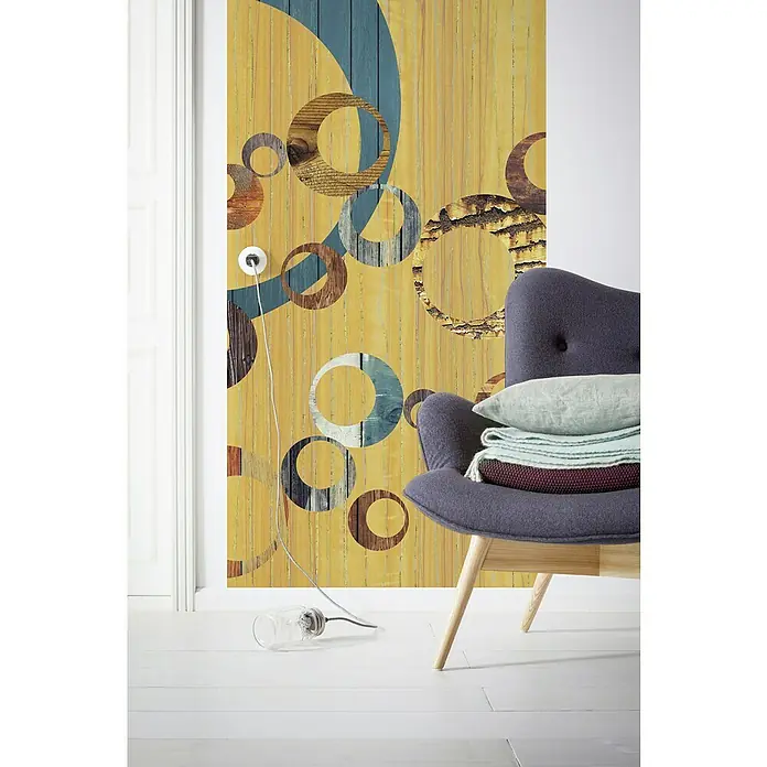 Komar Infinity Fototapete United Panel - 1 -tlg., B x H: 100 x 250 cm, Vlies - Wandbild mit gelbem Holzhintergrund und bunten Kreisen, ergänzt durch einen Sessel mit Kissen und Decken, verleiht dem Raum einen modernen Touch.