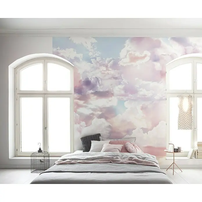 Komar Infinity Fototapete Clouds - 3 -tlg., B x H: 300 x 250 cm, Vlies - Schlafzimmer mit einem Bett, das mit grauer Bettwäsche, Kissen und einer rosa Tagesdecke bezogen ist, vor einer Wandtapete mit Wolkenmotiv.