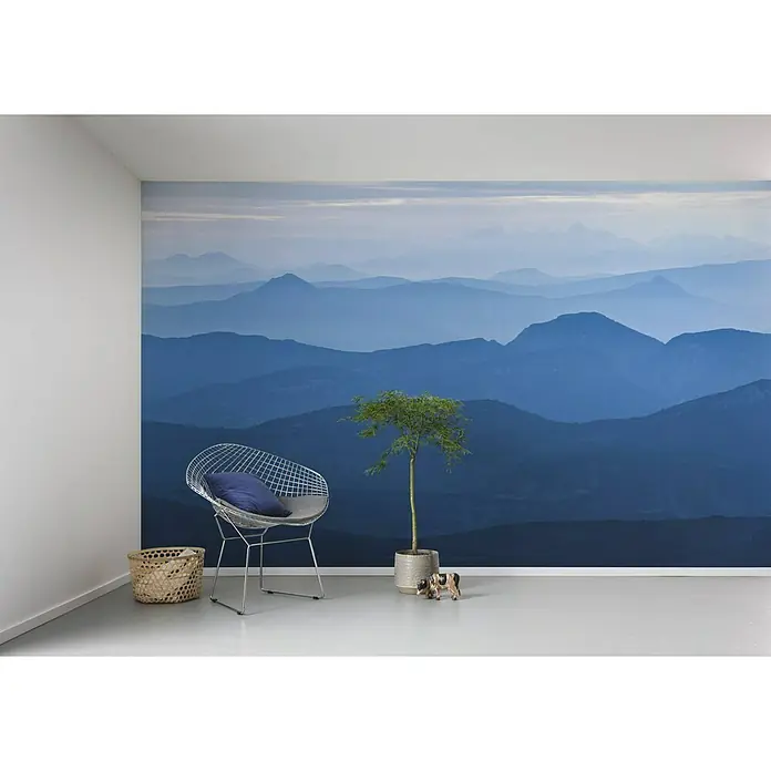 Komar Infinity Fototapete Blue Mountain - 4 -tlg., B x H: 400 x 250 cm, Vlies - Eine stilvolle Raumgestaltung mit einer Fototapete, die ein blaues Bergpanorama zeigt, einem Drahtsessel, Korb und einer Zimmerpflanze.