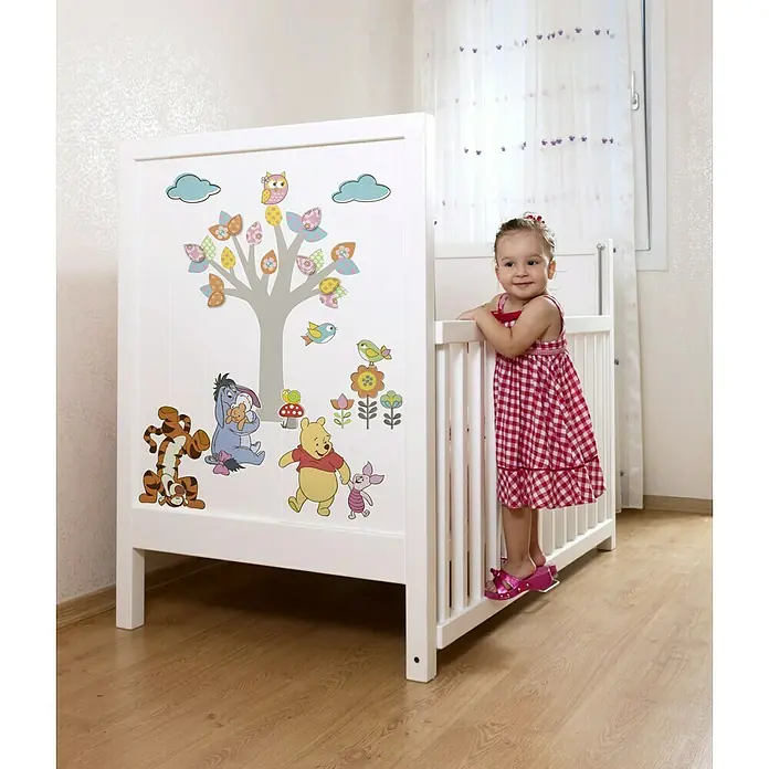 Komar Disney Edition 4 Dekosticker Winnie Pooh Nature Lovers - 14 -tlg., Bunt - Ein weißes Babybett mit Winnie-the-Pooh-Motiven und einem kleinen Mädchen, das sich an der Seite des Betts anlehnt.