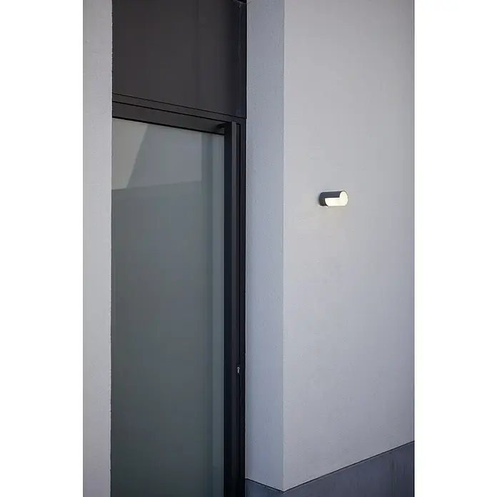 Lutec Plafón LED redondo Cyra - Blanco cálido, IP54 - Primer plano de una lámpara de pared gris junto a una ventana, que emite un brillo suave y acogedor.