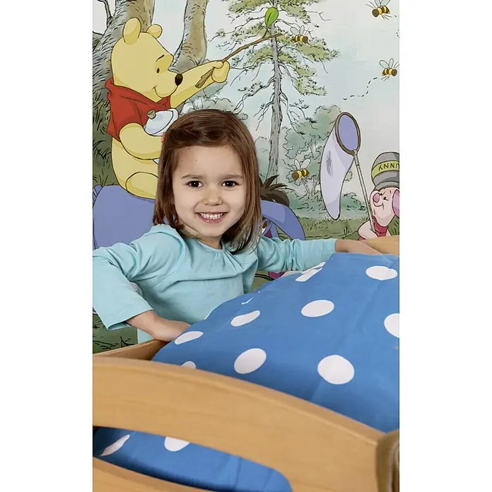 Komar Disney Edition 4 Fototapete Pooh´s House - 4 -tlg., B x H: 368 x 127 cm, Papier - Ein lächelndes Mädchen steht vor ihrem Kinderbett mit blauem Bettbezug mit weißen Punkten vor einer Winnie Puuh-Tapete.