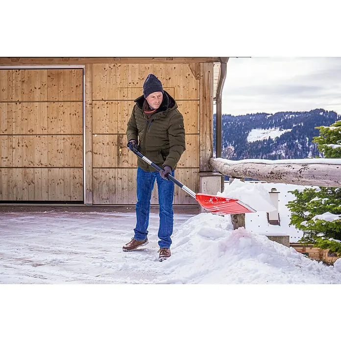 Freund Victoria Schaufel 67305 - 36 cm, Rot - Mann schaufelt Schnee mit einer roten Schneeschaufel vor einem Holzgebäude. Winterliche Szene mit Bergen im Hintergrund.