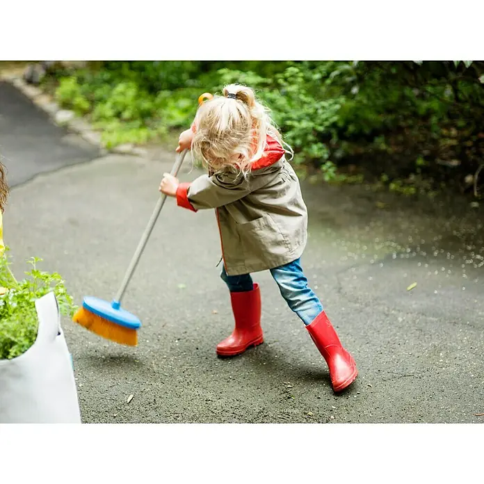Fiskars Kinder-Besen My First - Arbeitsbreite: 26 cm, Kurzstiel - Ein kleines Mädchen in roten Gummistiefeln fegt mit einem Besen eine Auffahrt im Freien. Das Kind trägt eine Jacke und Jeans.
