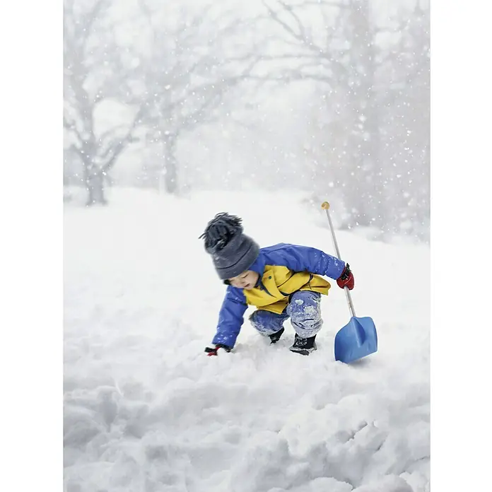 Fiskars Kinder-Schaufel My First - Art Stiel: Kurzstiel - Ein Kind in Winterkleidung spielt im Schnee mit einer blauen Schneeschaufel, während es stark schneit und Bäume im Hintergrund zu sehen sind.