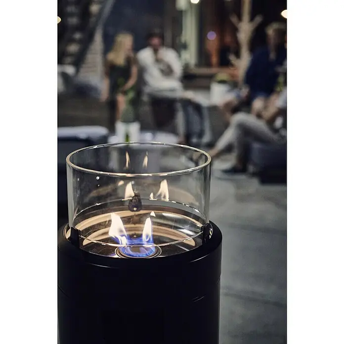 Enders Feuerstelle Nova - Ø x H: 36 x 102 cm, Schwarz - Nahaufnahme eines kleinen Tischkamins mit Glasaufsatz und blauer Flamme, umgeben von verschwommenen Personen im Hintergrund.