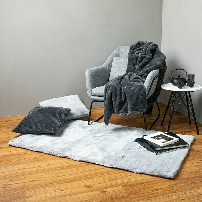 Decke HappyGraphit, 200 x 150 cm, 100 % Polyester - Einrichtungsszene mit einem flauschigen Teppich, einem grauen Sessel mit einer Decke, Kissen und einem Beistelltisch mit Teegeschirr.