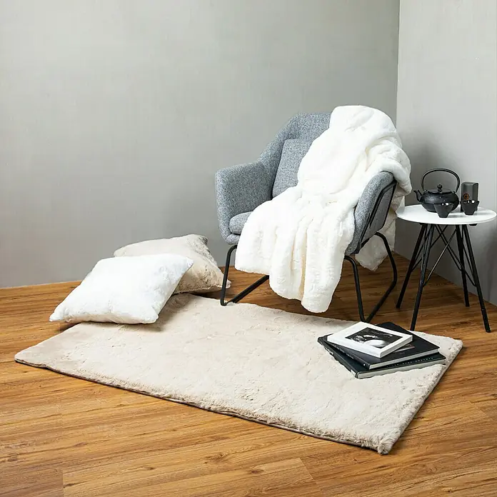 Decke HappyElfenbein, 200 x 150 cm, 100 % Polyester - Ein weicher, rechteckiger Teppich in sanftem Beige liegt auf einem Holzboden, neben einem Sessel und einem Beistelltisch.