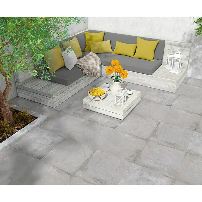 BHS Showroom Pavimento porcelánico Origin - 60 x 60 cm, Acero - Acogedor patio con baldosas de piedra gris, una cómoda zona de estar con cojines amarillos y una mesa de madera blanca con flores y limones.