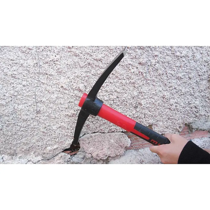 Peddinghaus Kreuzhacke - Länge: 37,5 cm, Stahl - Eine Hand hält eine rot-schwarze Spitzhacke, die eine strukturierte weiße Wand aufbricht und rote Ziegel freilegt.