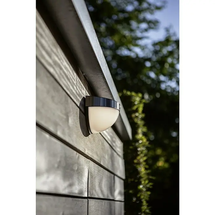 Lutec Aplique solar LED para exterior con sensor - 1,2 W, Acero inoxidable, Blanco neutro, Plateado - Una lámpara de pared de metal y vidrio montada en una pared de madera, que emite un brillo suave. Tiene un aspecto moderno y elegante.