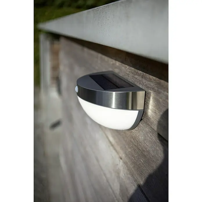 Lutec Aplique solar LED para exterior con sensor - 1,2 W, Acero inoxidable, Blanco neutro, Plateado - Primer plano de una lámpara de pared solar con sensor de movimiento montada en una pared de madera, que proporciona iluminación exterior eficiente.