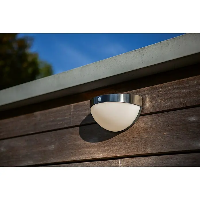 Lutec Aplique solar LED para exterior con sensor - 1,2 W, Acero inoxidable, Blanco neutro, Plateado - Primer plano de una lámpara redonda de exterior con sensor de movimiento montada en una pared de madera.