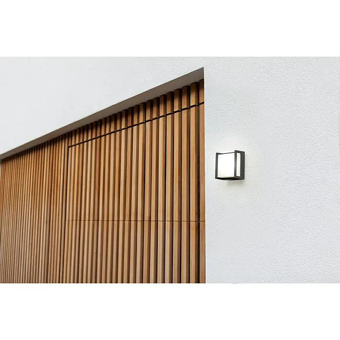 Lutec Aplique exterior LED Qubo - 8 W, Antracita, IP54 - Una lámpara de pared moderna en forma de cubo negro, montada en una pared blanca junto a un panel de madera, proporcionando iluminación exterior elegante.