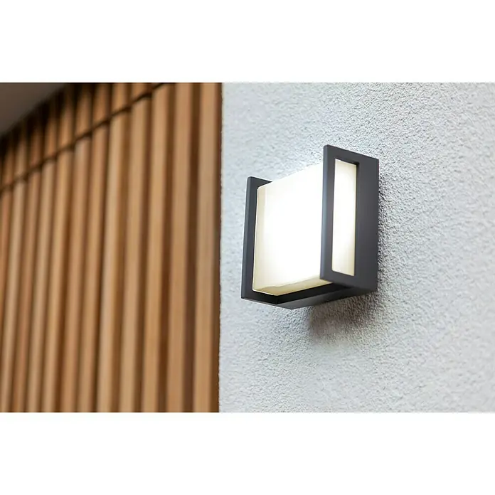 Lutec Aplique exterior LED Qubo - 8 W, Antracita, IP54 - Un aplique de pared cuadrado moderno con marco negro y luz difusa, montado en una pared con textura para una iluminación elegante y cálida.