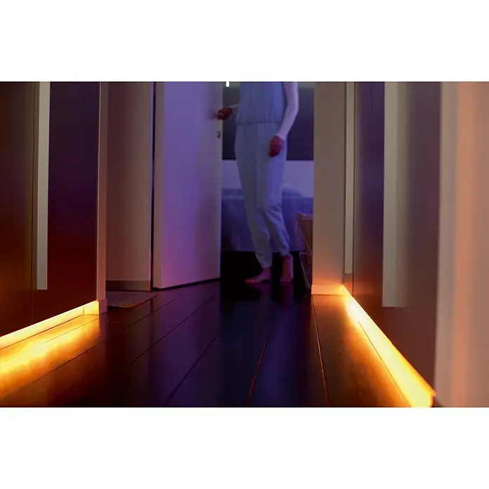 Philips Hue LED-Band Plus Erweiterung - 1 m, RGBW - Warmes, sanftes LED-Licht erhellt einen Flur mit dunklem Holzboden und schafft eine gemütliche Atmosphäre.