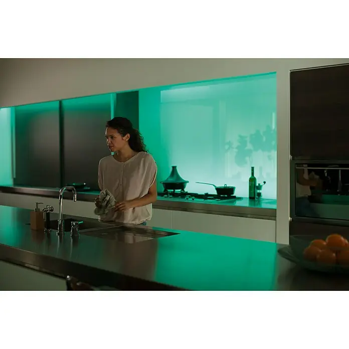 Philips Hue LED-Band Plus Erweiterung - 1 m, RGBW - Frau steht an einer modernen Spüle, indirektes Licht beleuchtet die Küche. Minimalistisches Design mit grünem Farbton für eine entspannende Atmosphäre.