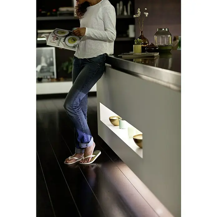 Philips Hue LED-Band Plus Erweiterung - 1 m, RGBW - Eine Frau in Jeans und Flip-Flops liest eine Zeitschrift in einer modernen Küche mit stilvoller Beleuchtung und Dekoration.