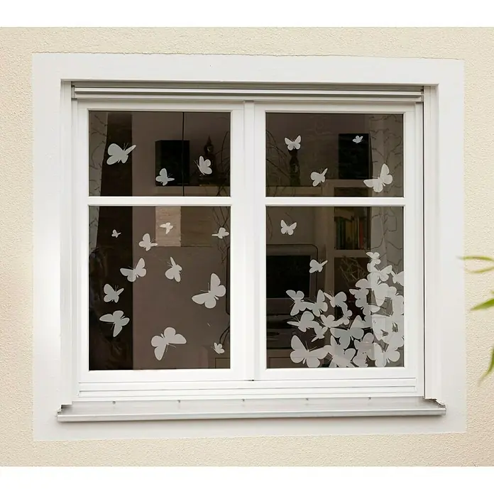 Komar Fenstersticker Schmetterlinge - Weiß, 31 x 31 cm, 21 -tlg. - Das Bild zeigt eine Fensterdekoration mit weißen Schmetterlingsmotiven, die auf die Glasscheiben aufgebracht wurden. Die Fenster sind weiß umrandet und mit einem Rollladen versehen.