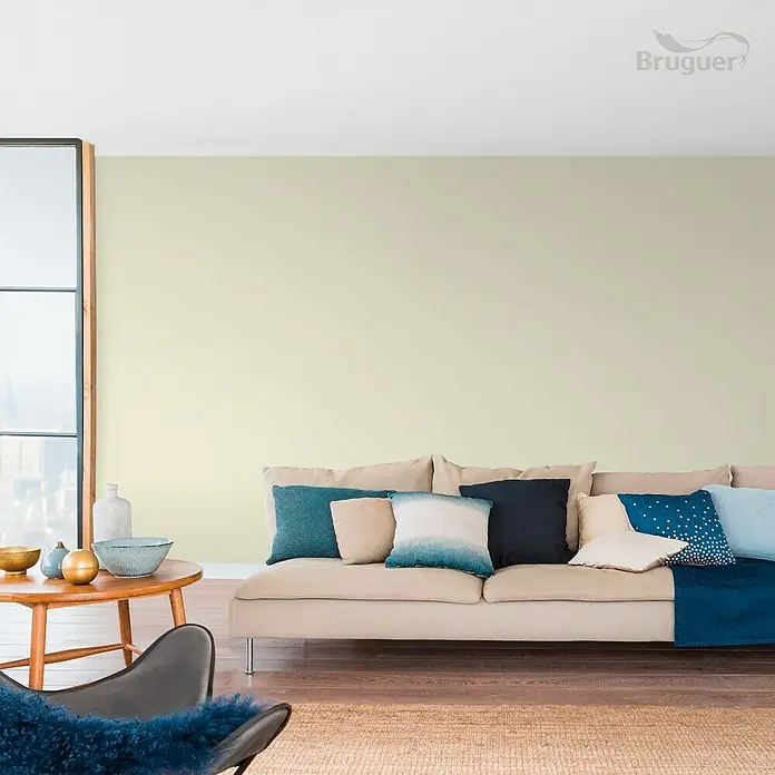 Bruguer Colores del Mundo Tester de pintura - Amazonas verde suave, Mate, 30 ml - Sala de estar luminosa con sofá beige, cojines azules y una pequeña mesa de madera con cuencos decorativos. La pared está pintada en un suave tono amarillo.