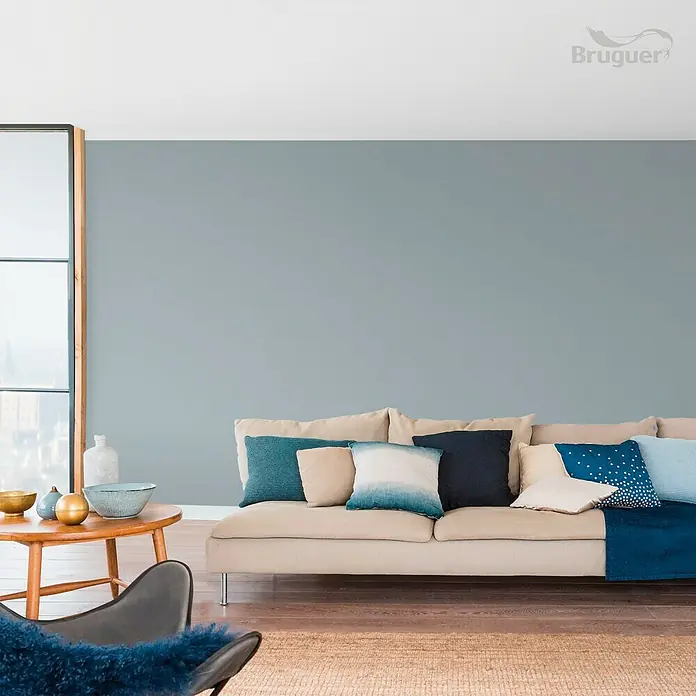 Bruguer Colores del Mundo Tester de pintura - Escandinavia gris intermedio, Mate, 30 ml - Elegante sala de estar con sofá beige, cojines azules y mesa de madera. La pared está pintada en un tono azul relajante, creando un ambiente tranquilo.