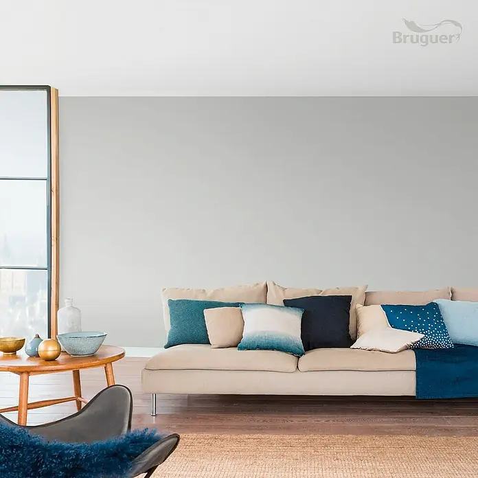 Bruguer Colores del Mundo Tester de pintura - Escandinavia matiz de gris, Mate, 30 ml - Salón luminoso con sofá beige y cojines azules. La pared está pintada de un gris suave, complementando el interior moderno y la marca Bruguer.