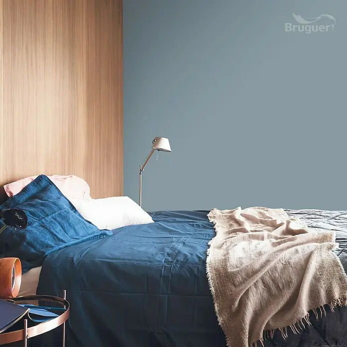 Bruguer Colores del Mundo Tester de pintura - Escandinavia gris natural, Mate, 30 ml - Dormitorio con pared pintada de azul, cama acogedora y revestimiento de madera. Logotipo de Bruguer en la esquina.
