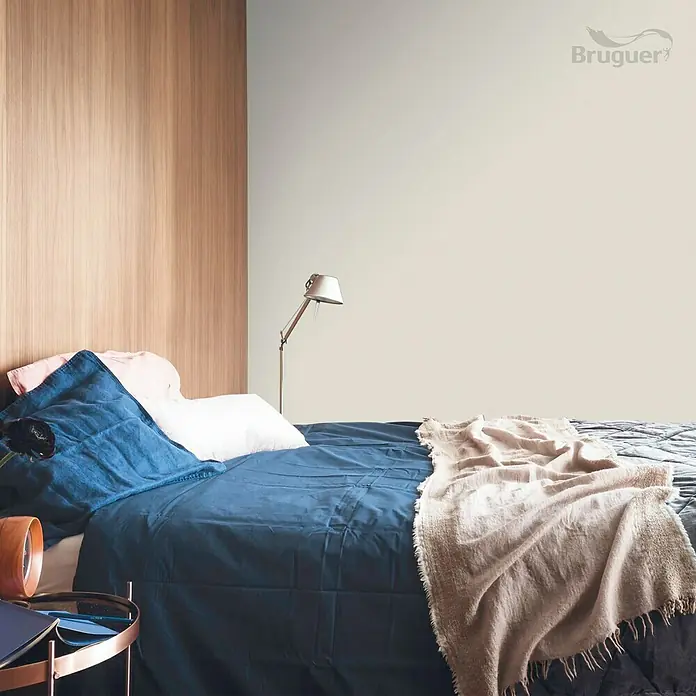 Bruguer Colores del Mundo Tester de pintura - Nepal matiz de beige, Mate, 30 ml - Dormitorio con ropa de cama azul, manta clara y pared de madera. La pared está pintada en un tono suave. Logotipo de Bruguer en la esquina.