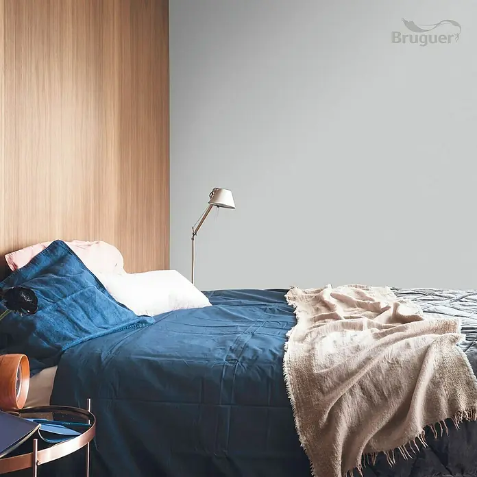 Bruguer Colores del Mundo Tester de pintura - Escandinavia matiz de gris, Mate, 30 ml - Dormitorio con ropa de cama azul y pared de madera. El logo de Bruguer es visible en la parte superior derecha. Una lámpara está en la mesa de noche.