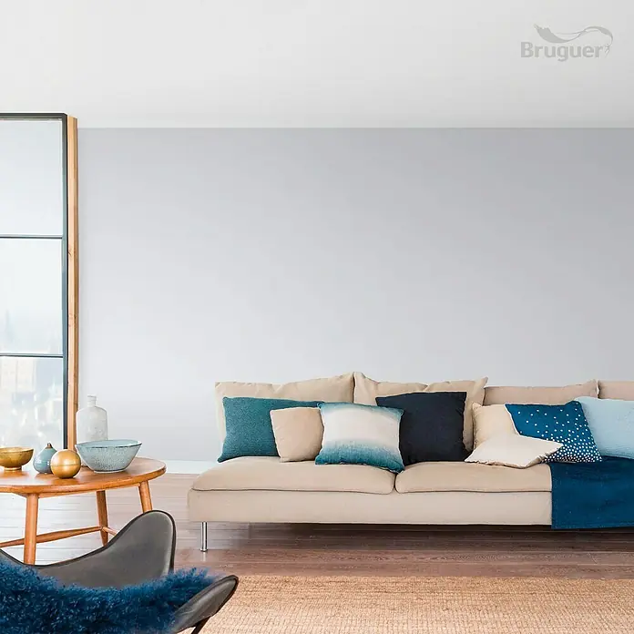 Bruguer Colores del Mundo Tester de pintura - Mediterráneo matiz de lila, Mate, 30 ml - Acogedora sala de estar con sofá beige, cojines azules y mesa de madera. El color claro de la pared y la alfombra crean un ambiente acogedor.