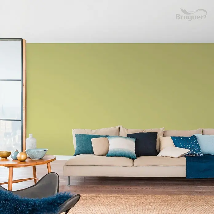 Bruguer Colores del Mundo Tester de pintura - Amazonas verde natural, Mate, 30 ml - La imagen muestra un interior moderno con una pared verde pintada con Bruguer, un sofá cómodo y cojines decorativos. La habitación parece acogedora.