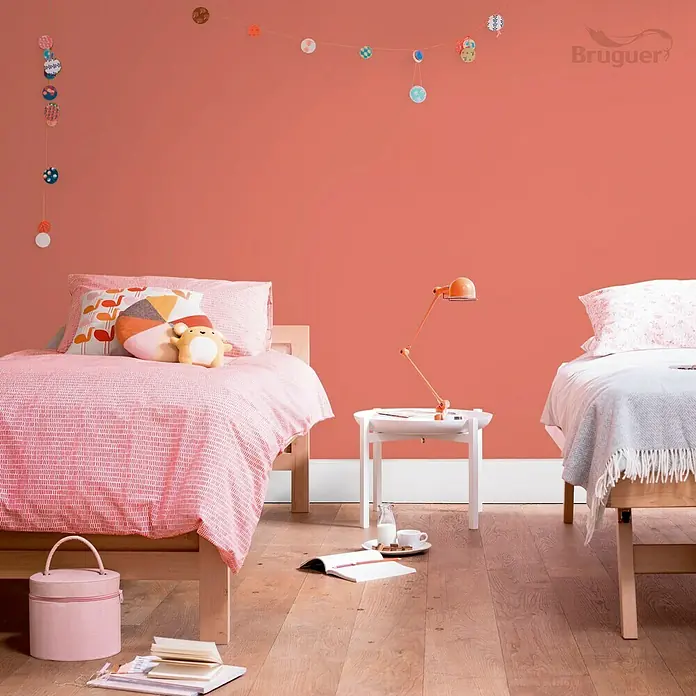 Bruguer Ultra Resist Tester de pintura - Rojo carácter, Mate, 30 ml - Dos camas infantiles en una habitación luminosa con paredes de color coral, decoradas con guirnaldas. Una mesita de noche con una lámpara entre las camas.